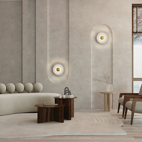 A minimalist living room showcases a cream curved sofa and white rug. Two Fiore Dimmable LED Circle Disc Wall Lights in brass adorn the textured beige wall. Wooden tables and a chair enhance the modern decor, with sunlight filtering through blinds for an elegant touch.