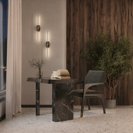 A stylish room showcases a brass and black marble table with an open book, a vase with twigs, and a black chair. Two Renaissance LED Wall Lights illuminate the wooden panel wall. A tall potted plant adds greenery beside a large window with sheer curtains on the left.