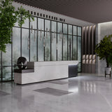 A modern lobby with minimalist design features a marble reception desk, geometric sculpture, and Ordo LED Short Pendant Lights with smoky cylindrical glass. Large smoky glass windows frame the space adorned with indoor plants, while a polished stone floor leads to sculptural wall decor.