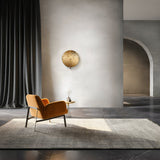 In a modern interior, a mustard yellow chair sits on a large gray rug, facing a small round wooden table with a vase. A textured wall decor piece hangs near the Mare Wall Light - Gold With Round Mirrored Glass Diffuser. Tall dark curtains and an arched doorway complete the elegant ambiance.