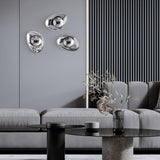 A modern living room showcases a sleek gray sofa and black glass coffee table. Three abstract silver sculptures decorate the gray slatted wall, alongside the elegant Mabell Wall Light - Chrome. A vase with dried flowers sits on the table, enhancing the contemporary decor.