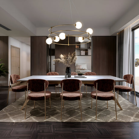 A modern dining room showcases a large rectangular table surrounded by eight velvet chairs on a geometric rug, illuminated by the Balance 6 Light Suspension - Gold With Opal Globe chandelier. A vase with pampas grass adorns the table, while floor-to-ceiling windows flood the space with natural light.