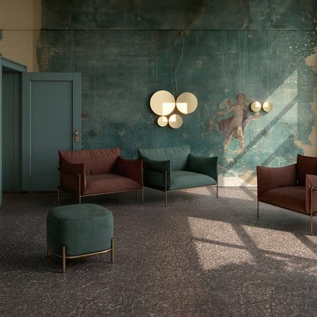 A chic seating area includes two brown armchairs, a green ottoman, and a teal wall with abstract art. Jupiter 10W 3000K LED Wall Lamps in brushed brass offer soft lighting, as sunlight streams through the window, casting shadows on the terrazzo floor.