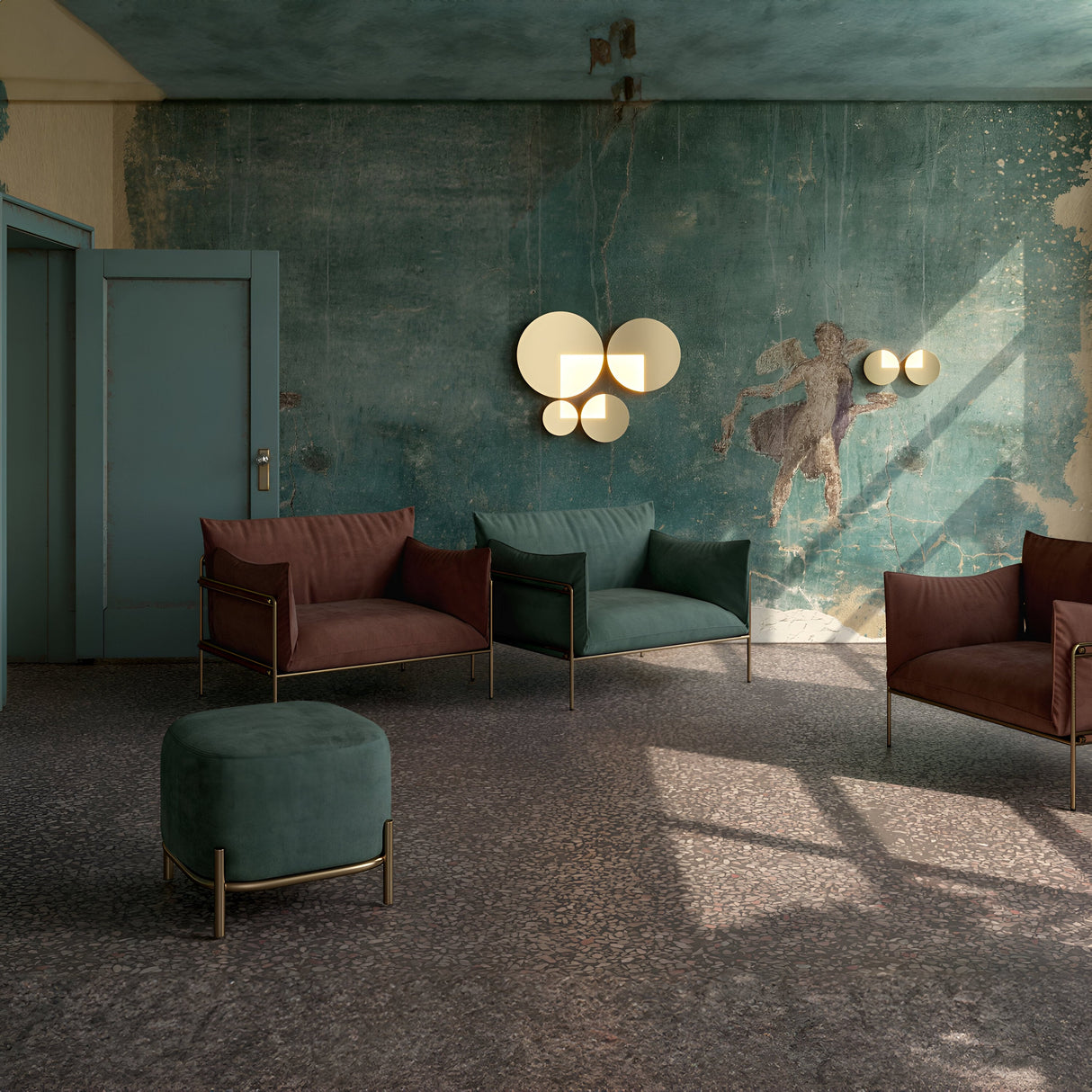 A vintage-style room with faded teal walls and modern furniture, including brown and green armchairs, features a matching green ottoman. The space is illuminated by the Jupiter LED Wall Light 12W 3000K in brass, with light streaming through large windows.