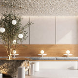 A modern interior with a natural design theme features a large rock table, cushioned benches, the Basic Form Table Lamp in gold with white frosted glass, small round tables with globe lamps, and a leafy tree. Beige and wood tones dominate the decor, enhanced by a textured ceiling.