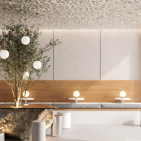 A modern interior with a natural design theme features a large rock table, cushioned benches, the Basic Form Table Lamp in gold with white frosted glass, small round tables with globe lamps, and a leafy tree. Beige and wood tones dominate the decor, enhanced by a textured ceiling.