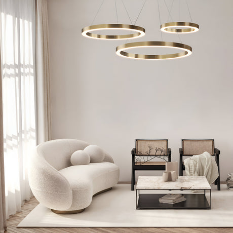 A modern living room boasts a curvy white sofa, two beige cushioned chairs, and a marble-topped coffee table. Three Saturno LED Circular Suspension Lights in brass hang overhead, while a large window with sheer white curtains optimizes the natural light.