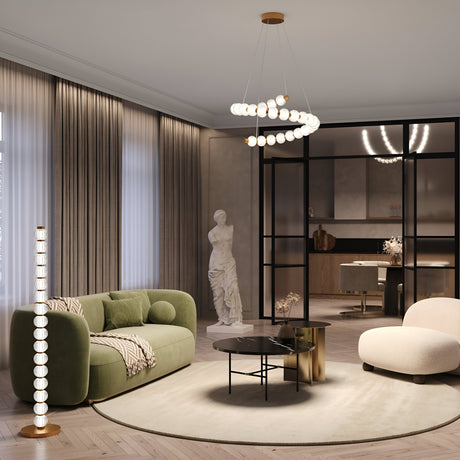 A modern living room showcases a green sofa, white armchair, and circular coffee table. Decorative lighting includes the Amulet LED Spiral Suspension Light - Gold With Ribbed Glasses. A spiral chandelier brightens the golden-framed room and a statue sits by floor-to-ceiling curtains.