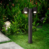 The Wall Street 2 Light Bollard 55cm in black stands elegantly on a lush green lawn, surrounded by tropical plants and stones. This modern outdoor lamp enhances the gardens ambiance as a stone pathway meanders through, elevating the lighting experience.