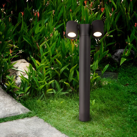 The Wall Street 2 Light Bollard 55cm in black stands elegantly on a lush green lawn, surrounded by tropical plants and stones. This modern outdoor lamp enhances the gardens ambiance as a stone pathway meanders through, elevating the lighting experience.