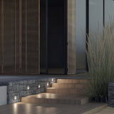 A modern exterior entrance showcases stone steps with IP54 protection, highlighted by the Bil Outdoor LED Recessed Wall Light in white for safety and style. Tall ornamental grasses and wooden sliding panels partially cover a glass wall on the left for a sleek, contemporary look.