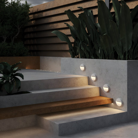 Concrete steps featuring the Mane Outdoor LED Round Wall Light - White guide you to a terrace adorned with large green plants and a wooden privacy screen. The energy-efficient LED lighting softly enhances the space, creating a modern, serene ambiance that embodies contemporary aesthetics.