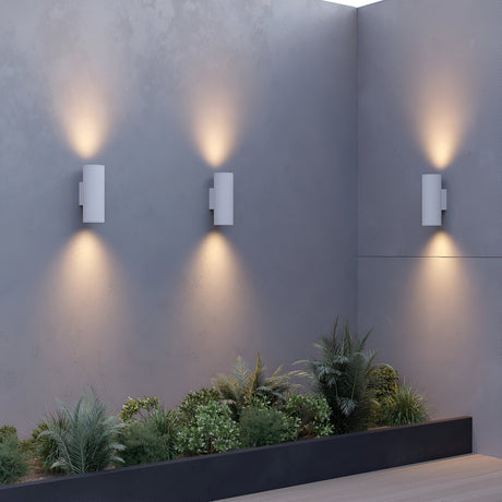 A modern outdoor area showcases three Shim Outdoor Wall Lights 24W 3000K IP65, emitting a warm glow against a gray wall. Below the LEDs, a planter box with assorted greenery adds nature to the minimalist design.