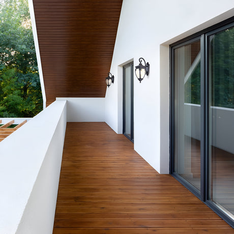 A modern balcony showcases a sloped wooden ceiling and floor. Two glass sliding doors on the right are flanked by Fleur Outdoor Wall Light 60W in bronze, casting a warm glow. The white walls contrast beautifully with natural surroundings, enhanced by stylish bronze-finish accents.