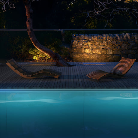 In a nighttime poolside scene, two wooden lounge chairs sit on a deck. The Edge Outdoor Landscape LED Ground Light - Black softly illuminates the water, casting a serene glow. A tree and stone wall in the background enhance the tranquil ambiance of this weather-resistant setting.