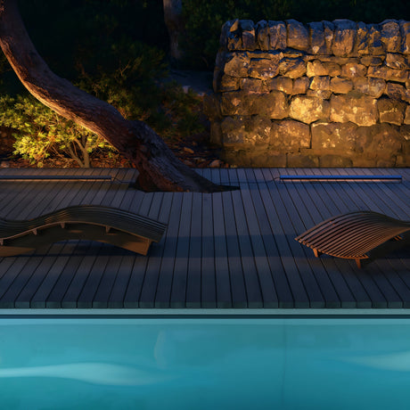 A tranquil poolside scene features two wooden lounge chairs on a deck, with the softly glowing blue pool water complemented by a 3000K LED lamp. A tree and stone wall are gently lit by the graphite-colored Edge Outdoor Landscape LED Ground Light, creating a cozy ambiance.