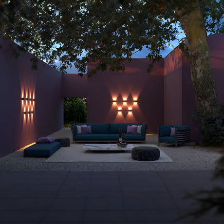 Outdoor patio with purple walls, modern teal sofa, chairs, and low table. Strato Outdoor LED Wall Light creates geometric patterns. A large tree provides partial shade as the evening sky looms above.