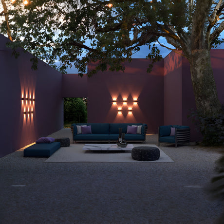 The outdoor lounge, framed by purple walls and Strato Outdoor LED Wall Lights (6W 3000K - Grey), radiates a warm glow. Blue seating surrounds a low white table, shaded by a large tree, as the IP54-rated fixtures stylishly illuminate the space with corrosion-resistant elegance at dusk.