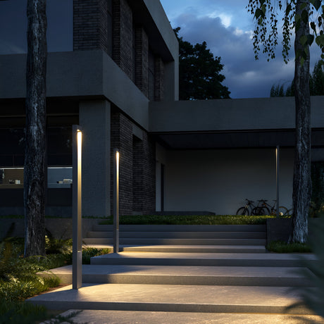 At dusk, the modern house exterior is lit by Bonn Bollard Light 200cm in Graphite with integrated LED modules. Trees and plants encircle the area, bicycles rest near the entrance, and the darkening sky adds to the serene evening enhanced by this stylish outdoor lighting.