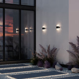 Rom Outdoor LED Up & Down Wall Lights illuminate a building exterior at dusk, their reflections visible in large glass doors. Ornamental plants and gravel decorate the ground below, with the Graphite finish anchoring the serene contemporary aesthetic.