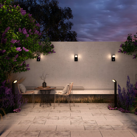 A serene outdoor patio features a wooden table with a chair and bench amidst lavender plants. Modern wall lights and Stock LED Bollard Light 3000K 7W - Graphite illuminate the concrete wall and tiled flooring. Twilight sky visible with silhouetted trees in the background.