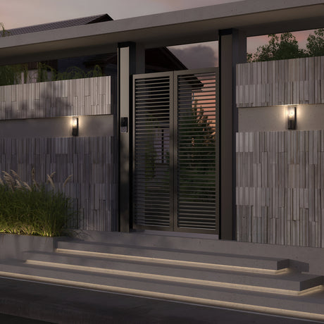 A modern front gate with horizontal slats is flanked by concrete walls featuring vertical tile patterns. Two Conf Outdoor Square Wall Lights, in a sleek graphite finish and boasting an IP65 rating, illuminate the entrance. Steps lead up to the gate with greenery on the left, enhancing the evening mood.