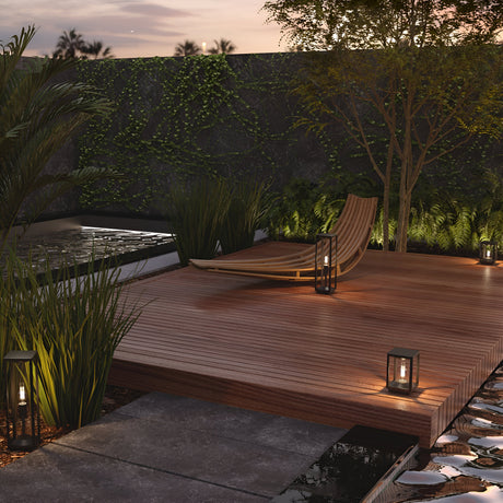 An outdoor deck features a wooden lounge chair amid lush greenery, softly illuminated by Cell Pedestal Light 22cm in Graphite. A serene water feature flows nearby, with ivy-covered walls under a twilight sky providing a tranquil backdrop.