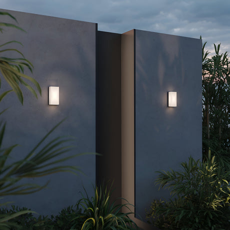 The Badd Outdoor Square Wall Light 3000K in graphite illuminates the textured concrete walls of a building at dusk. Energy-efficient LEDs highlight lush greenery, casting soft shadows and creating a serene atmosphere.