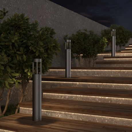 Outdoor wooden steps with built-in strip lights ascend alongside a stone wall, bordered by lush bushes. Vint LED Bollard Lights (3000K 9W - Graphite, 60cm) line the pathway under the night sky.