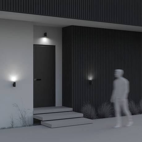 A dark rectangular front door with a three-step entrance is highlighted by the Bowery Outdoor 1 Light Wall Light - Grey (O574WL-01GR), casting soft glows. A blurred figure walks on the path lined with sparse grass in front of this minimalistic house.