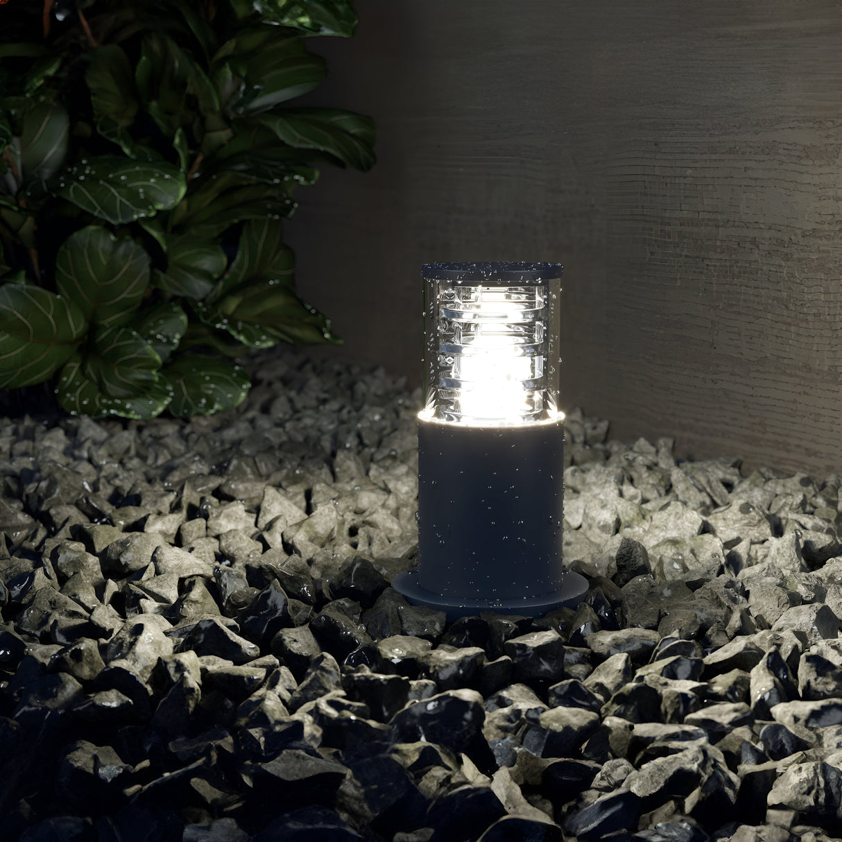 A Bronx Bollard Light 250mm - Black illuminates dark stones, highlighting garden pathways. Green leafy plants can be seen on the left, with a textured wall serving as a horizontal backdrop.