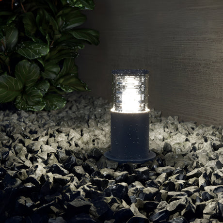A Bronx Bollard Light 250mm - Black illuminates dark stones, highlighting garden pathways. Green leafy plants can be seen on the left, with a textured wall serving as a horizontal backdrop.
