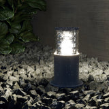 The Bronx Bollard Light 250mm - Grey stands on gray stones, casting a bright glow that illuminates glossy surfaces. Its corrosion-resistant design ensures durability, complemented by green foliage in the background for a natural touch.
