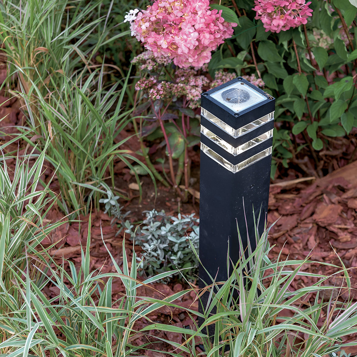 The Unter den Linden Bollard Light in black, featuring silver accents, is corrosion-resistant and stands vertically amidst lush greenery and pink flowers on brown mulch, providing subtle illumination to the garden.