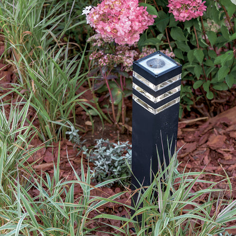 The Unter den Linden Bollard Light in black, featuring silver accents, is corrosion-resistant and stands vertically amidst lush greenery and pink flowers on brown mulch, providing subtle illumination to the garden.