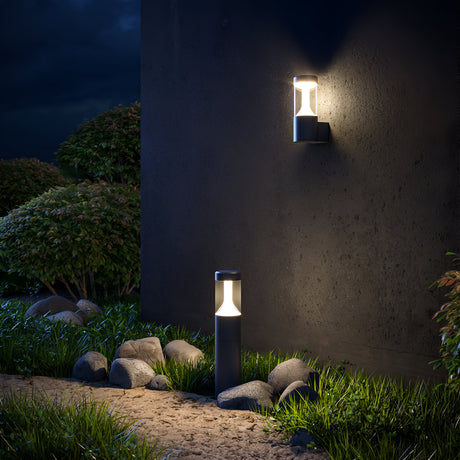 A nighttime garden showcases a modern wall lantern and a Koln LED Bollard Light 65cm - Black, with an LED module pathway light illuminating a stone path and lush green foliage against the dark sky.