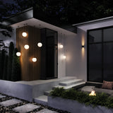 A modern house exterior at night showcases Erda Outdoor Pendant Lights in Black & White, with their spherical design illuminating a wooden-paneled entrance. Steps descend to a patio bordered by evergreens, featuring shrubs and a small fire pit—ideal for relaxing outdoor spaces.