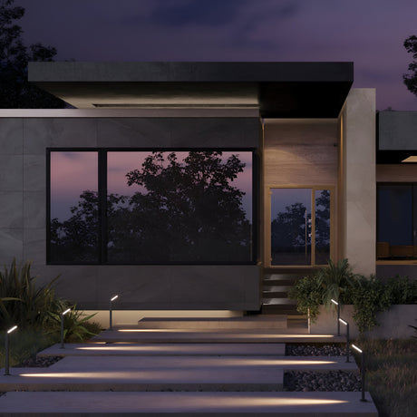 At dusk, a modern house with large windows and a flat roof sits under silhouetted trees against a purple and pink sky. The path to the entrance is lined with energy-efficient Paso LED Bollard Light 80cm - Black, enhancing the serene ambiance with subtle outdoor lighting.