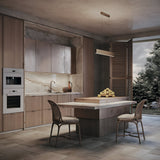 A modern kitchen features wooden cabinets, marble countertops, and a matching island with two wicker chairs. Atop the island rests a pile of apples. The Step LED Linear Suspension Light 4000K 30W - Gold efficiently illuminates the space, complemented by natural light from a large window overlooking the lush garden.