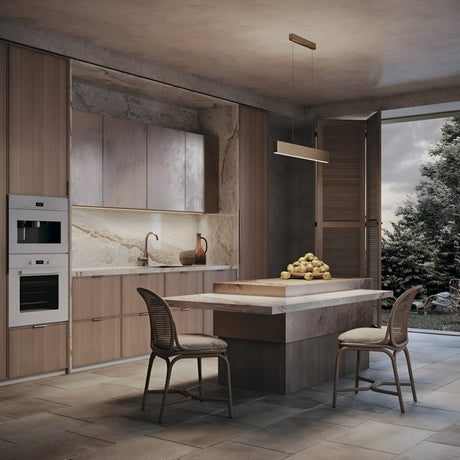 A modern kitchen features wooden cabinets, marble countertops, and a matching island with two wicker chairs. Atop the island rests a pile of apples. The Step LED Linear Suspension Light 4000K 30W - Gold efficiently illuminates the space, complemented by natural light from a large window overlooking the lush garden.