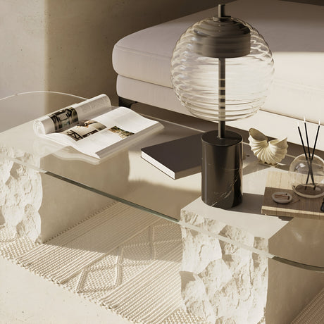 A modern glass coffee table with stone supports features an open book, a closed black book, the Mystic LED Table Lamp - Black With Marble Base, a seashell, and a reed diffuser. A cream sofa rests on a woven rug in the background, creating a cozy, minimalistic living space.