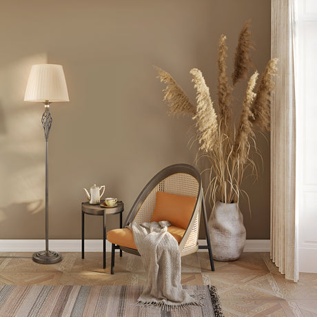 A cozy interior features a stylish chair with a soft beige blanket, the Grace Floor Lamp - Bronze with a pleated fabric shade, and a small round table holding a teapot and cup. Tall pampas grass in a vase stands beside sheer-curtained windows.