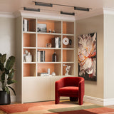 A cozy room features a large bookshelf with books and decor, a red velvet chair illuminated by a Points Rot LED Swivel Linear Track Light in matte black. A colorful floral painting adorns the wall, while a large potted plant rests on a patterned rug in the corner.