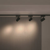 A modern interior with ceiling-mounted Vuoro 3-Circuit Trinity Track LED Spotlights in black (26W, 3000K) showcases a vertical slatted gray wall. These high CRI lights create a warm ambiance, highlighting the walls texture.