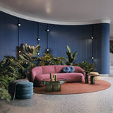 A modern living room includes a pink curved sofa with pillows on a circular brown rug, surrounded by a green glass coffee table and cylindrical side table. The blue textured wall features plants, round lights, and the Rado CCT LED Dimmable Magnetic Exility Track Spotlight for adjustable ambiance.