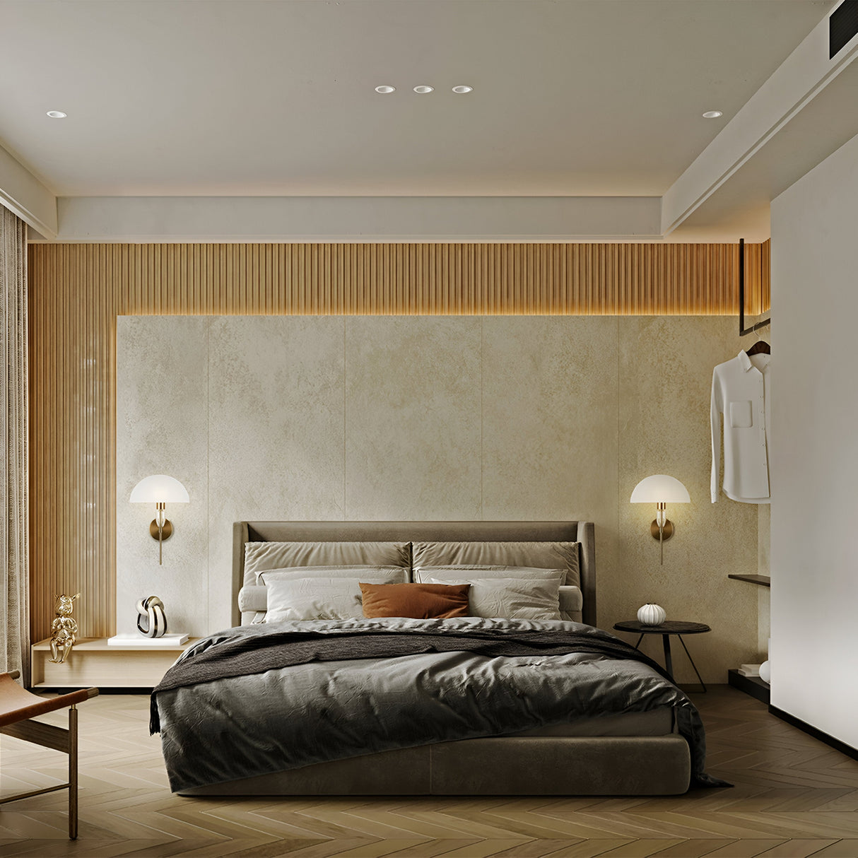 A modern bedroom with a textured accent wall showcases a large bed with gray and brown bedding. Two Prima Wall Lights - Brass with White Shade enhance the charm on either side. A wooden bench rests at the beds foot, and a white shirt hangs nearby. Warm lighting creates a cozy ambiance.