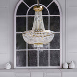 A Bella 8 Light Crystal Chandelier - Gold hangs against an arched window with dark glass panes. The room has white walls and a decorative white shelf adorned with two small white sculptures.