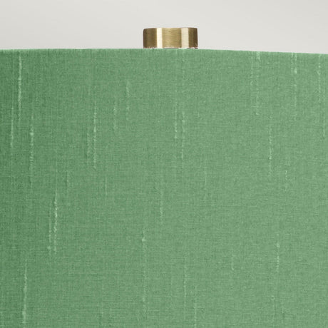 The Isla 1 Light Table Light - White/Green is captured in a close-up that highlights its textured green shade accented with subtle vertical stripes and topped with a small brass finial. Placed against a neutral backdrop, this lamp pairs elegantly with its white ceramic base, blending sophistication with an understated design.