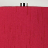 Close-up of the Isla 1 Light Table Light, showcasing a red fabric shade embellished with vertical thread patterns. Its white ceramic base harmonizes beautifully with the vibrant shade, and it is elegantly topped with a silver metal finial, all against a simple light gray background.