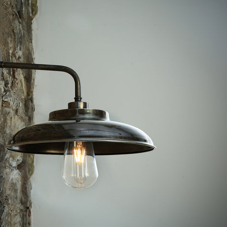 Islura Brass Wall Light IP65 with a wide, shallow disc shade on a swan-neck arm, exposed E27 bulb, mounted on a stone wall against a plain grey background.